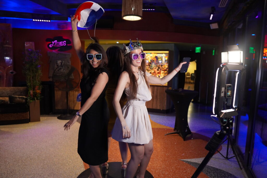 BOMBA Creative Co.'s luxury photo booth experience at a nightlife venue in Orlando featuring two guests enjoying fun photo props - bedazzled oversized sunglasses, a crown, and a colorful beach ball. The vibrant venue with colorful lighting showcases how our premium photo experiences create unforgettable moments at social events. #LuxuryPhotoBooth #OrlandoNightlifeEvents #PhotoBoothProps