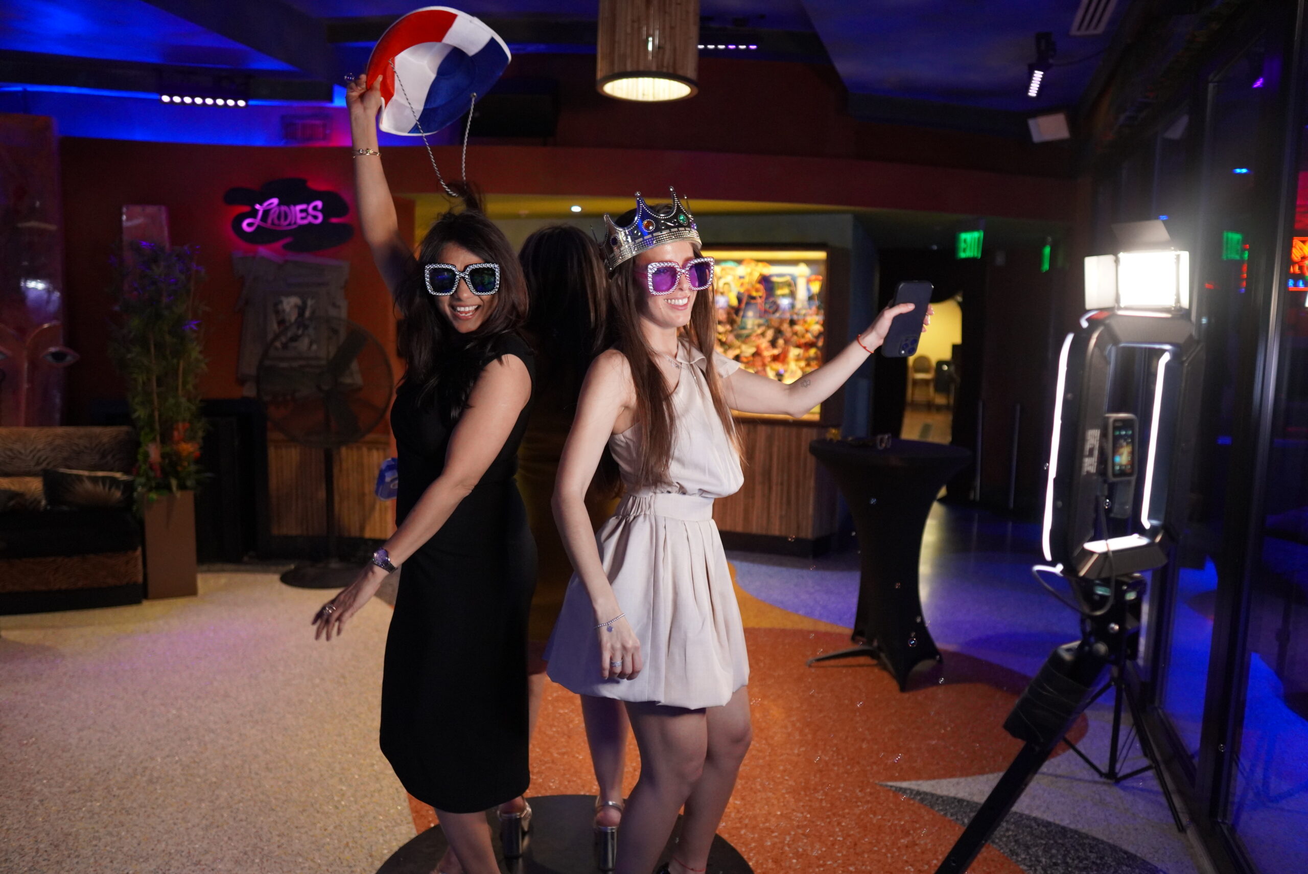 BOMBA Creative Co.'s luxury photo booth experience at a nightlife venue in Orlando featuring two guests enjoying fun photo props - bedazzled oversized sunglasses, a crown, and a colorful beach ball. The vibrant venue with colorful lighting showcases how our premium photo experiences create unforgettable moments at social events. #LuxuryPhotoBooth #OrlandoNightlifeEvents #PhotoBoothProps